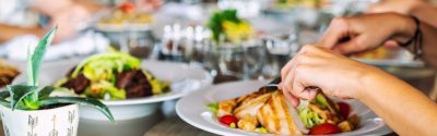 Two hands using a fork and knife to eat their meal with other plates of food in the background